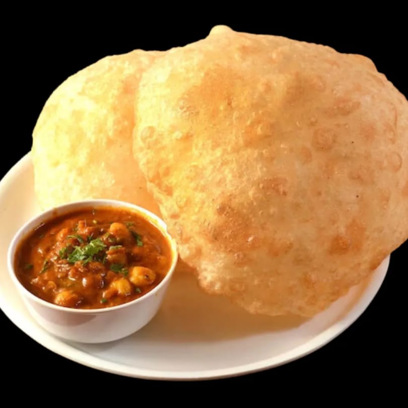 Two golden-brown Indian puri breads served with a small bowl of chana masala on a white plate.