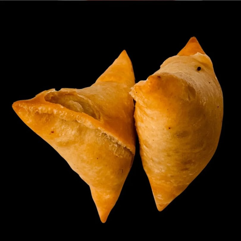 Two golden-brown, crispy samosas with a flaky crust, against a dark background.